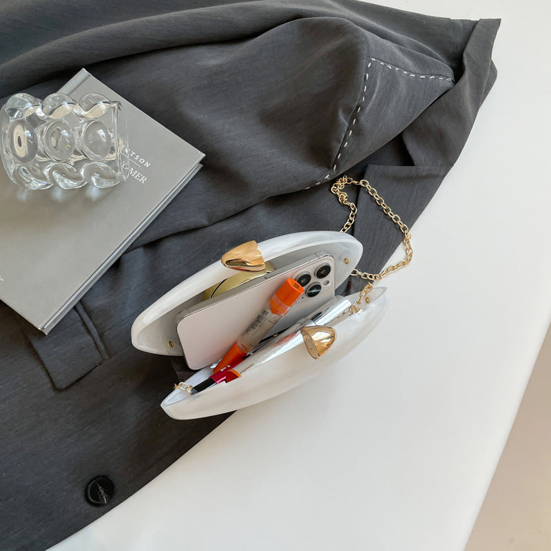 Oval shell acrylic evening bag opened, showcasing mobile phone and pens, stylishly placed on grey blazer.