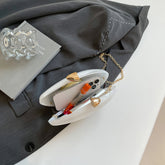 Oval shell acrylic evening bag opened, showcasing mobile phone and pens, stylishly placed on grey blazer.